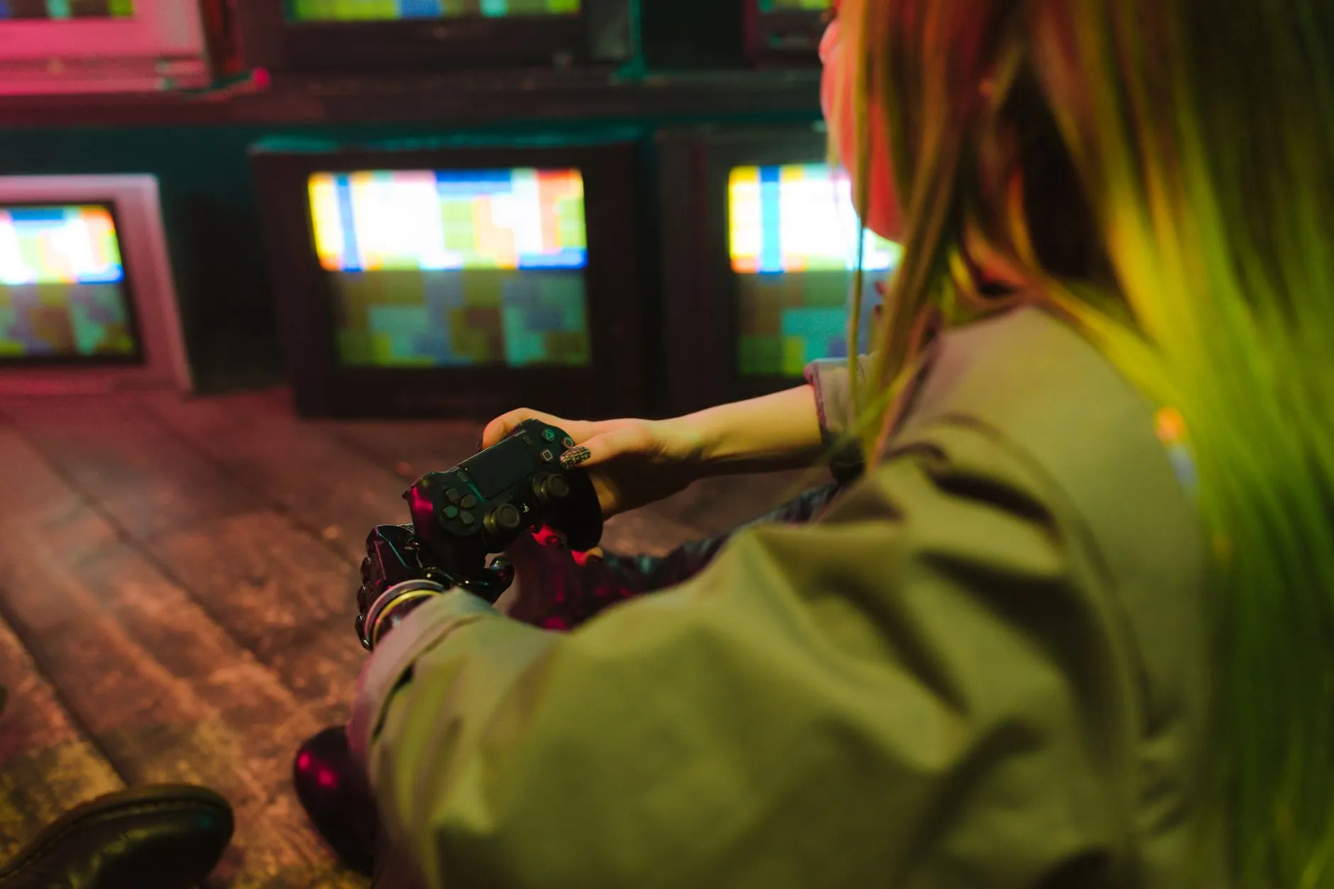 A player with green hair sits on a wooden floor holding a game controller, facing a wall of glowing CRT televisions.