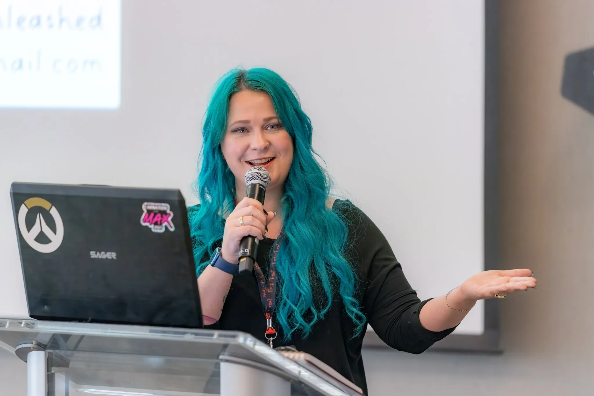 Tara, with long teal hair, speaks into a handheld microphone at a laptop podium and gestures to the audience.